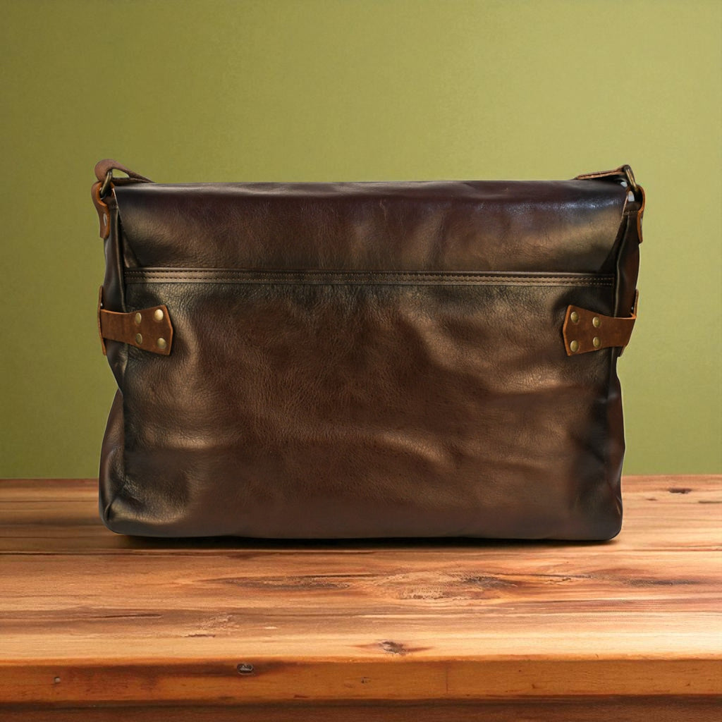 Brown leather messenger bag with gold accents on a wooden surface against a green wall
