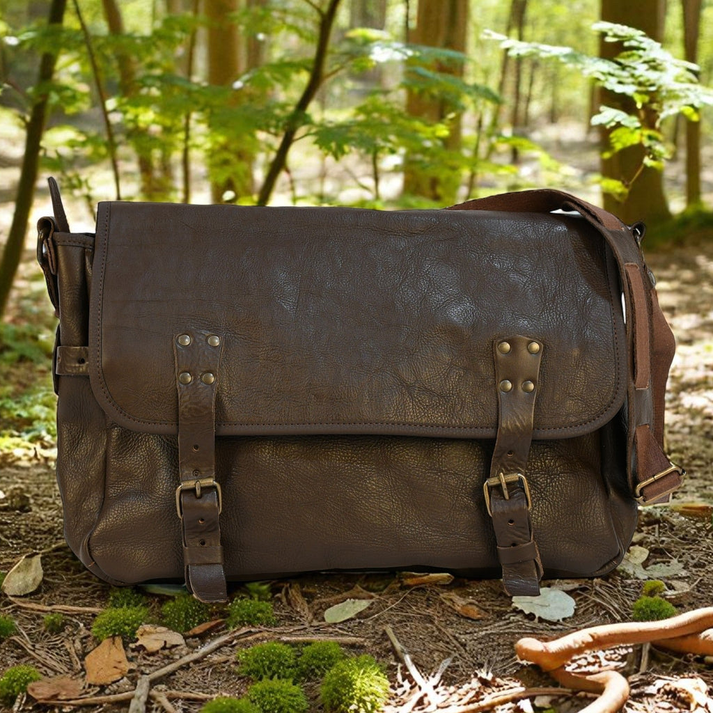 Brown leather modern rugged messenger bag on a forest floor with trees and foliage in the background