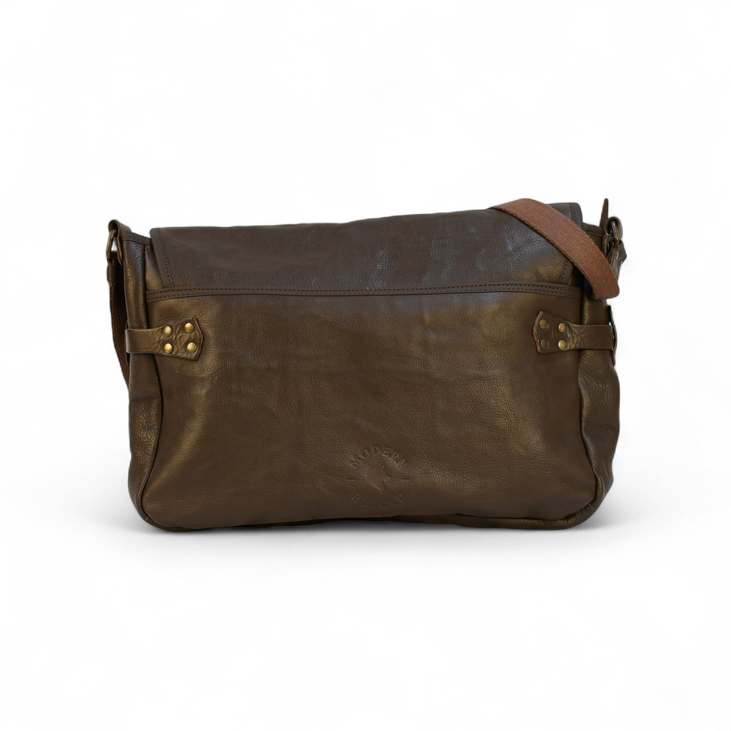 Brown leather modern rugged messenger bag on a forest floor with trees and foliage in the background