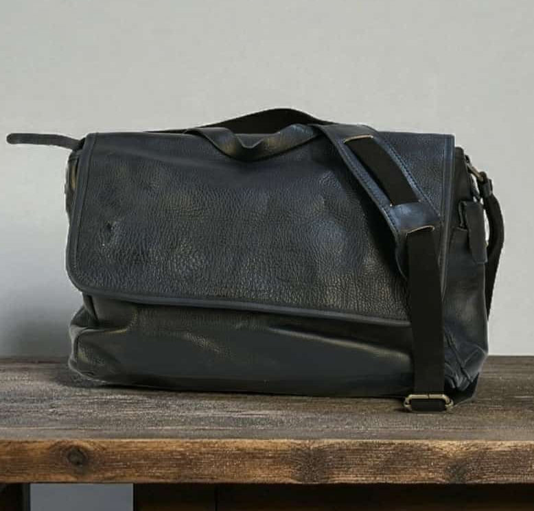 Black leather modern rugged messenger bag on a wooden table with a gray background