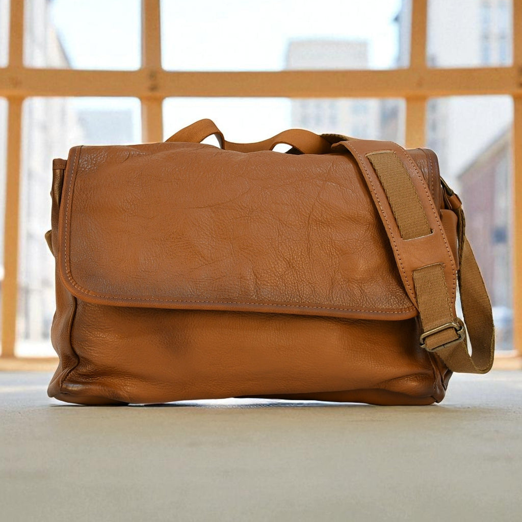 Brown leather modern rugged messenger bag on a surface with a window in the background