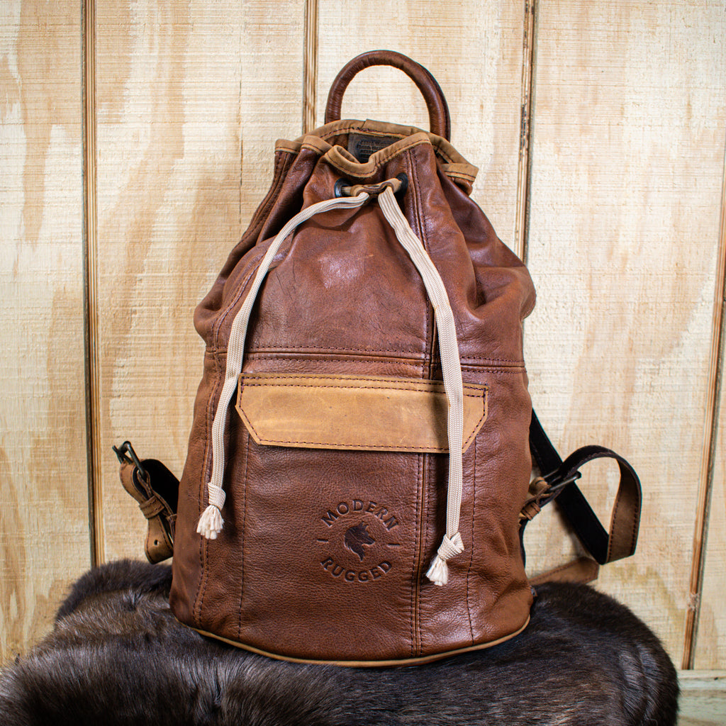 A brown leather bucket bag with brass zippers and buckles, placed upright with a wooden background. The bag has an adjustable strap and appears to have multiple compartments.