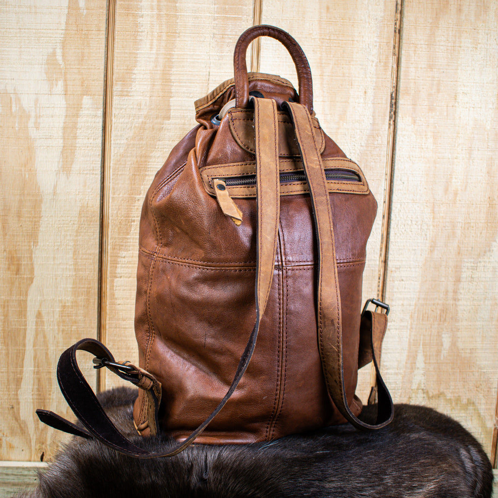 A brown leather bucket bag with brass zippers and buckles, placed upright with a wooden background. The bag has an adjustable strap and appears to have multiple compartments.