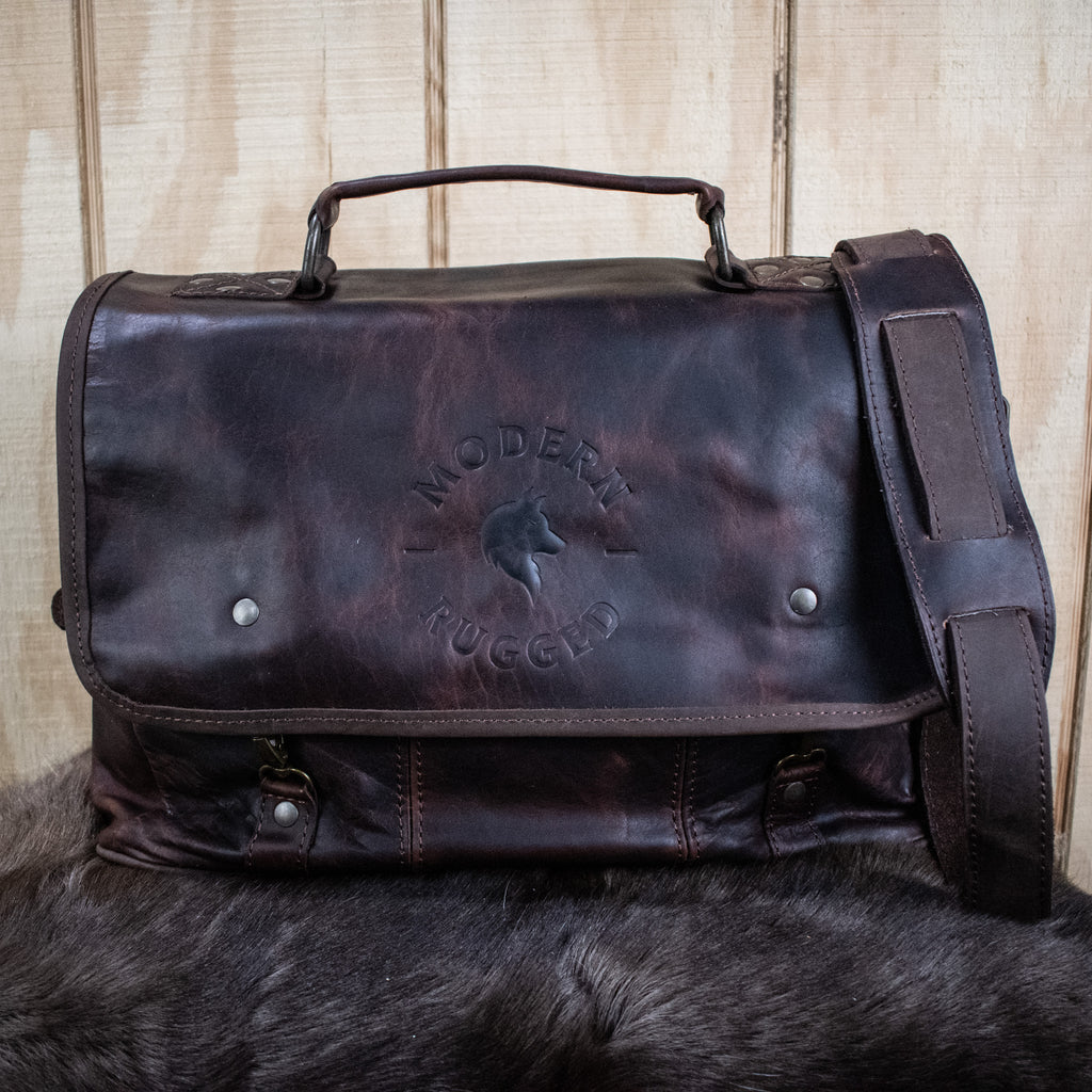 A brown leather laptop bag with brass zippers and clasps, featuring a side pouch and an adjustable strap, placed on a furry surface.