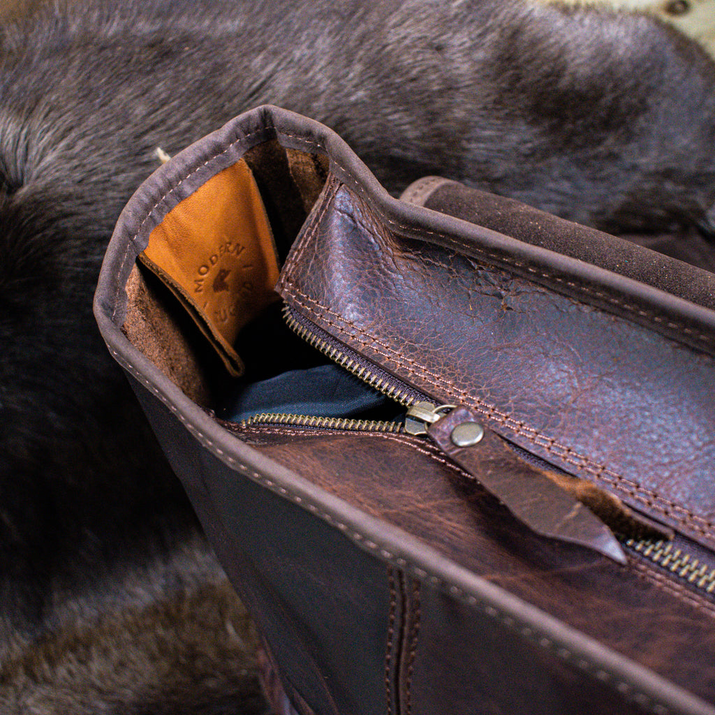 A brown leather laptop bag with brass zippers and clasps, featuring a side pouch and an adjustable strap, placed on a furry surface.